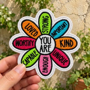 May include: A flower-shaped embroidered patch with a white center reading YOU ARE in black text. Surrounding petals feature the words LOVED, STRONG, IMPORTANT, KIND, UNIQUE, ENOUGH, CAPABLE, and WORTHY in black text against orange, lime, blue, orange, pink, pink, lime, and magenta backgrounds.