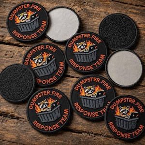 May include: A collection of circular embroidered patches featuring a raccoon sitting in a dumpster with a laptop surrounded by flames. The text DUMPSTER FIRE RESPONSE TEAM is stitched in orange around the border. Some patches show the black hook-and-loop backing.