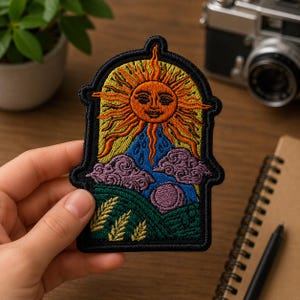 May include: An embroidered patch featuring an arched frame containing a stylized orange sun with a face, radiating yellow rays. Below the sun are blue raindrops, purple clouds, a green landscape with wheat stalks, and a purple circular moon.