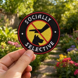 May include: A circular embroidered patch with a black border and a red outer ring. The center features a yellow background with a black silhouette of a dog and a pointing hand, crossed by a red diagonal line. The text reads SOCIALLY SELECTIVE.