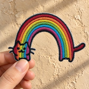 May include: An embroidered patch featuring a cat with a rainbow-colored body and head, trailing a long, arched rainbow tail. The design uses vibrant red, orange, yellow, green, blue, and purple threads with a thick black border outlining the cat shape.