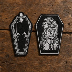May include: Two embroidered patches with black and white thread. The left patch is coffin-shaped, featuring a vampire figure and a small skull. The right patch is a hexagonal shape displaying a detailed portrait of a historical figure with long hair.