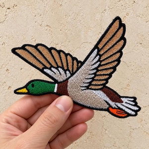 May include: An embroidered patch featuring a mallard duck in flight with wings spread wide. The duck has a vibrant green head, a yellow beak, brown and tan wings with white accents, and a bright orange tail tip, all outlined in black thread.