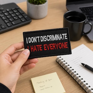 May include: A rectangular black embroidered patch with white text that reads I DON'T DISCRIMINATE and red text that reads I HATE EVERYONE. The patch features a clean, stitched border and is held against a desk setting with office supplies.