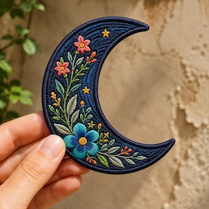 May include: A crescent moon-shaped embroidered patch featuring a dark blue background with a large blue flower at the base, smaller pink flowers, green leaves, and tiny yellow stars stitched along the curved surface of the fabric.