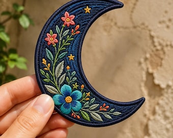 May include: A crescent moon-shaped embroidered patch featuring a dark blue background with a large blue flower at the base, smaller pink flowers, green leaves, and tiny yellow stars stitched along the curved surface of the fabric.