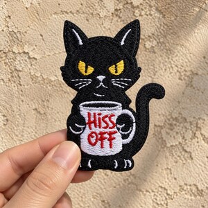 May include: An embroidered patch featuring a black cat with yellow eyes holding a white mug. The mug has the red text Hiss Off printed on the front. The cat has a grumpy expression with white whiskers and a curved tail extending to the right side.