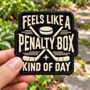 May include: A black embroidered patch with cream-colored text that reads FEELS LIKE A PENALTY BOX KIND OF DAY. The design features a central hockey puck and two crossed hockey sticks with a small four-pointed star below the puck.