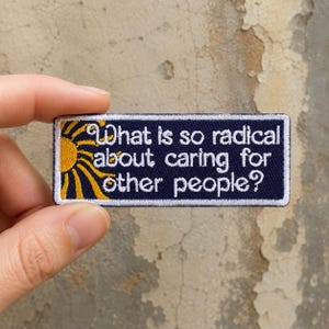 May include: A rectangular embroidered patch with a dark blue background and a white border. It features a yellow sun graphic on the left and the white embroidered text "What is so radical about caring for other people?" centered on the patch.