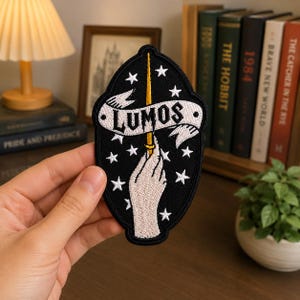 May include: An oval-shaped embroidered patch with a black background featuring a hand holding a yellow wand pointing upward. A white banner across the center displays the text "LUMOS" in black letters, surrounded by small white embroidered stars.