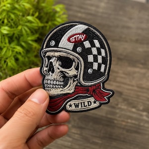 May include: An embroidered patch featuring a skull wearing a black and white checkered motorcycle helmet. The helmet has a red oval with the text STAY. A red bandana is tied around the skull's neck, and a banner below reads WILD with stars.