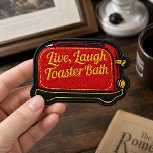May include: A red embroidered patch shaped like a toaster with a black border and yellow stitching. The text "Live, Laugh Toaster Bath" is embroidered in yellow across the center of the toaster. The design includes yellow details for the lever.