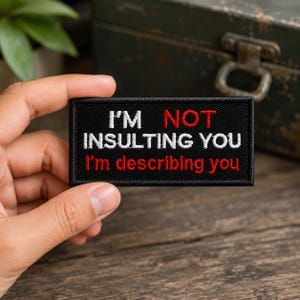 May include: A rectangular black embroidered patch featuring white and red text that reads I'M NOT INSULTING YOU I'm describing you. The patch has a black stitched border and is designed for attachment to fabric items like clothing or bags.