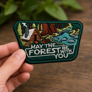 May include: A trapezoidal embroidered patch featuring a forest scene with a log cabin, a smoking chimney, a small campfire, and a blue stream. The text "MAY THE FOREST BE WITH YOU" is stitched in white thread against a dark green background.