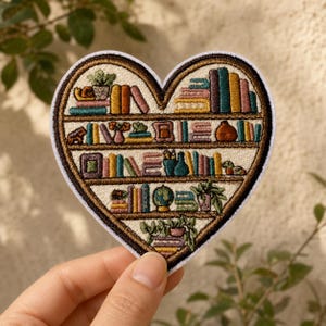 May include: A heart-shaped embroidered patch featuring a multi-tiered bookshelf design. The shelves are filled with colorful books, potted plants, a small globe, and decorative objects, all rendered in detailed thread work with a dark brown border.