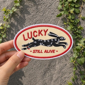 May include: Oval-shaped patch with a cream background, red border, and the word "LUCKY" in red script. A black and white rabbit is in the center, with the words "- STILL ALIVE -" below. The patch is held by a hand.