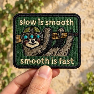 May include: A rectangular embroidered patch with a dark green background and a black border. It features a sloth wearing a tactical helmet with night vision goggles, holding a rifle. The text reads: slow is smooth, smooth is fast.
