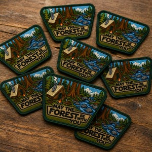 May include: A collection of trapezoidal embroidered patches featuring a forest scene with a cabin, a river, and a stone bridge. The text 'MAY THE FOREST BE WITH YOU' is printed in bold letters at the bottom of each patch with a dark green border.