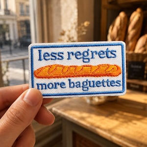 May include: A rectangular embroidered patch with a white background and a blue border. The text "less regrets" appears in blue above an orange baguette illustration, with the text "more baguettes" in blue below the bread graphic.