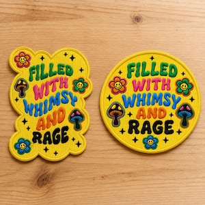 May include: Two embroidered patches with yellow backgrounds. One is cloud-shaped, the other circular. Both feature the text "FILLED WITH WHIMSY AND RAGE" in green, pink, blue, orange, and black, surrounded by colorful smiley flowers and mushrooms.