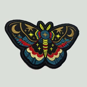 May include: An embroidered moth patch featuring a black base with vibrant red, blue, and yellow thread details. The design includes celestial motifs such as golden crescent moons and four-pointed stars, with a striped yellow and black body.