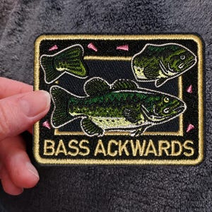 May include: A rectangular embroidered patch with a black background and gold border. It features a large green bass fish, a smaller bass head, and a tail fin. Small pink triangular accents are scattered around. The text BASS ACKWARDS is in gold.