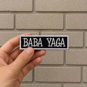 May include: A rectangular black patch with the embroidered text "BABA YAGA" in white, framed by a white border. The patch is held in a person's hand against a neutral brick background.