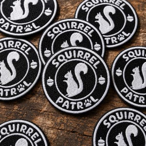May include: A collection of circular embroidered patches featuring a black background with a white squirrel silhouette in the center. Each patch displays the text SQUIRREL PATROL in white lettering, accompanied by small white acorn and paw print icons.