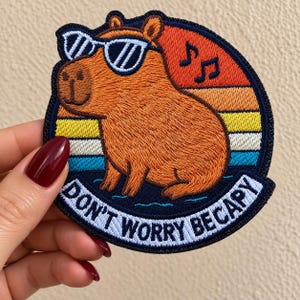 May include: An embroidered patch featuring a brown capybara wearing white sunglasses against a sunset background of orange, yellow, white, and blue stripes. Two musical notes are above the capybara, and the text DON'T WORRY BECAPY is below.