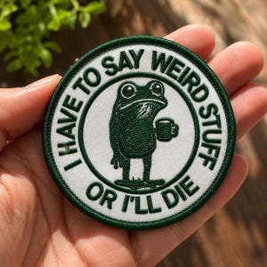 May include: A circular embroidered patch with a white center and a dark green border. The patch features a dark green illustration of a frog holding a mug, surrounded by the text "I HAVE TO SAY WEIRD STUFF OR I'LL DIE" in dark green capital letters.