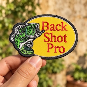 May include: An oval-shaped embroidered patch featuring a green and black fish graphic on the left side and the red text Back Shot Pro on a yellow background. The patch has a thick black border and is designed for attachment to clothing or gear.