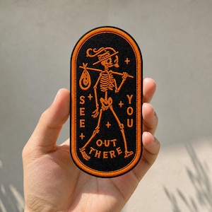 May include: An oval-shaped black patch with an orange embroidered border. The design features an orange skeleton walking while carrying a bindle on a stick. The text "SEE YOU OUT THERE" is embroidered in orange, accompanied by small star symbols.