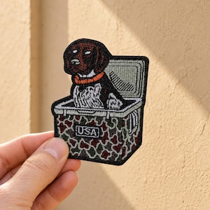 May include: An embroidered patch featuring a brown and white dog with an orange collar sitting inside a camouflage cooler. The cooler has a black border and the text USA printed in a white rectangular box on the front of the camouflage pattern.