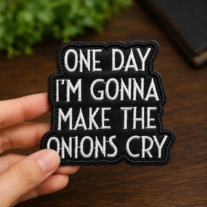 May include: A black embroidered patch with a scalloped border featuring the white stitched text ONE DAY I'M GONNA MAKE THE ONIONS CRY. The patch has a rectangular shape with rounded corners and a dense, textured fabric finish with clear white lettering.