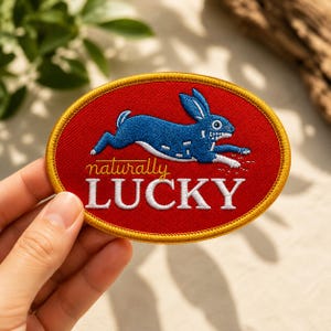 May include: An oval embroidered patch with a red background and a gold border. It features a blue rabbit in a running pose above the text "naturally LUCKY" in white and yellow script and block lettering. The design is centered on the patch.