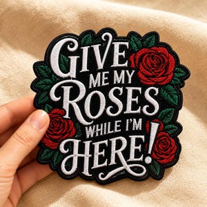 May include: A black embroidered patch featuring the white text GIVE ME MY ROSES WHILE I'M HERE! surrounded by red roses and green leaves. The patch has a scalloped edge and is designed for application to fabric items as a decorative accessory.