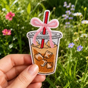 May include: An embroidered patch shaped like a plastic cup filled with iced coffee, featuring a red straw and a light pink bow tied around the top. The design includes brown liquid, white ice cubes, and a clear lid with a white border outline.