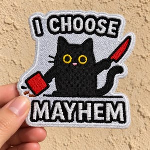 May include: A white embroidered patch featuring a black cat with yellow eyes. The cat holds a red knife in one paw and a red container in the other. The text "I CHOOSE" is embroidered in black above the cat, and "MAYHEM" is embroidered in black below.