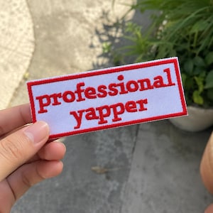 May include: A rectangular embroidered patch with a white background and a red border. The text "professional yapper" is stitched in red, lowercase, sans-serif letters across the center of the patch, creating a clear and bold visual design.