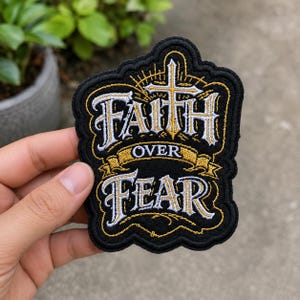 May include: A black embroidered patch featuring the text FAITH OVER FEAR in white and gold lettering. A gold cross sits above the word FAITH, with decorative gold rays radiating outward. The patch has a black border and a scalloped edge shape.