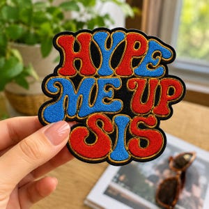 May include: An embroidered patch with a black background featuring the text HYPE ME UP SIS in a wavy, bold font. The letters are colored in alternating red and blue with a gold-colored border outlining each letter and the entire patch shape.