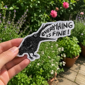 May include: An embroidered patch featuring a black bird with its beak wide open, accompanied by a white speech bubble containing the black embroidered text "EVERYTHING IS FINE!". The patch has a white border and a textured, stitched appearance.