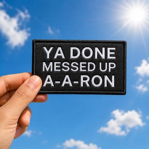 May include: A black rectangular patch with white embroidered text that reads "YA DONE MESSED UP A-A-RON". The patch is held against a bright blue sky with the sun shining in the background.