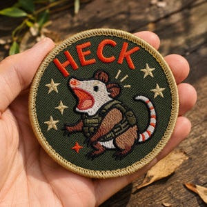 May include: A circular embroidered patch with a dark olive green background, featuring a brown and white opossum wearing a tactical vest. The word HECK is stitched in orange above the opossum, surrounded by several small embroidered stars.