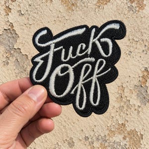 May include: A black embroidered patch featuring the white text "Fuck Off" in a cursive script font. The patch has a thick black border and a textured fabric surface, with the lettering standing out in a bright white thread against the black background.