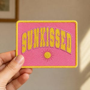 May include: A rectangular embroidered patch with a pink background and a yellow border. The word SUNKISSED is stitched in yellow with a pink outline in an arched font. Below the text is a small, circular yellow sun icon with radiating lines.