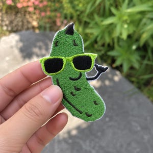 May include: An embroidered patch shaped like a green pickle wearing bright lime green sunglasses with black lenses. The pickle has a small black smile, a few dark green stitch details, and one arm raised in a shaka hand gesture sign.