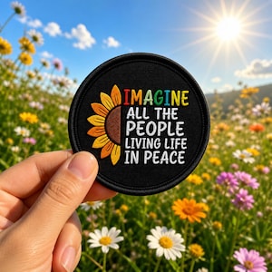 May include: A round black patch featuring a sunflower design and the words "IMAGINE ALL THE PEOPLE LIVING LIFE IN PEACE" in colorful text. The patch is held against a backdrop of a sunny field with wildflowers.