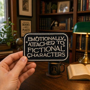 May include: A black embroidered patch with the text "Emotionally Attached to Fictional Characters" in cream-colored thread. The patch is rectangular with rounded corners and a cream border. The patch is held in front of a bookshelf and desk.