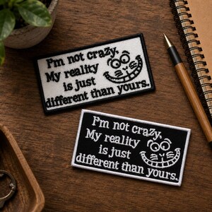 May include: Two rectangular embroidered patches featuring the text "I'm not crazy. My reality is just different than yours." and a stylized cat grin. One patch has a white background with black text, and the other has a black background with white text.